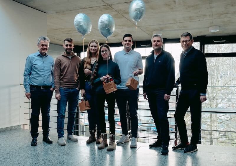 Gruppenfoto zum Ausbildungsabschluss 2025 bei der Firma Projekt-HLS in Weiden. Sieben Personen stehen in einem modernen Bürogebäude mit großen Fenstern. In der Mitte des Bildes sind drei Auszubildende zu sehen, die silberne Luftballons und kleine Geschenktüten in den Händen halten. Links und rechts von ihnen steht die Geschäftsführung des Unternehmens. Alle Personen lächeln und blicken in die Kamera.